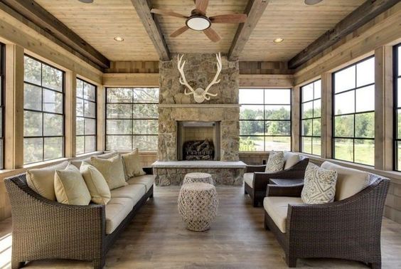 Vue intérieure d'une véranda en bois décorée d'une cheminée en pierres, avec un parquet en bois et des canapés et fauteuils qui se font face.