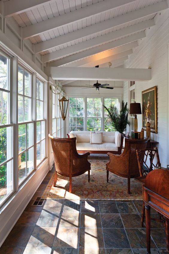 Vue intérieure d'une véranda en bois blanc décorée dans un style vintage, avec des fauteuils en rotin, un tapis et des meubles en bois couleur acajou.