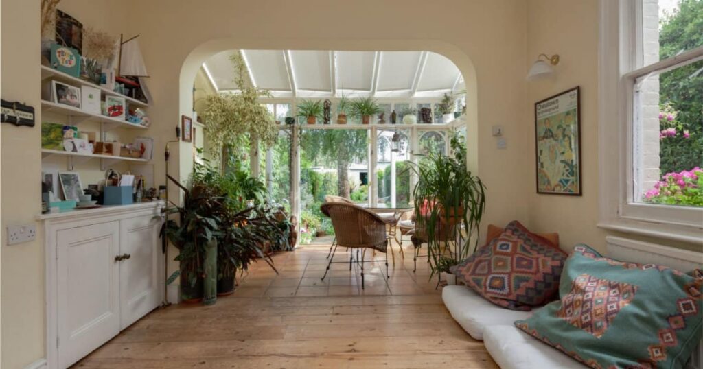 Vue intérieure d'une maison dans une pièce de détente avec des coussins par terre, qui mène à une véranda en bois décorée de plantes. 