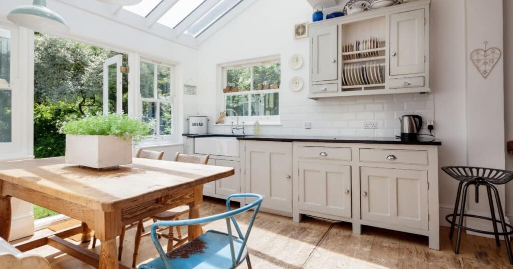 Vue intérieure d'une véranda en bois peinte en blanc et aménagée en cuisine, avec des plans de travail blancs en fond et une grande table en premier plan. 