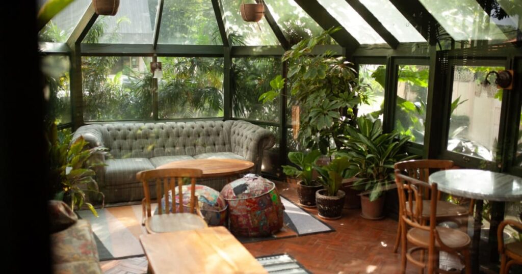 Vue intérieure d'une véranda victorienne aménagée en salon avec deux petites tables, des chaises et un canapé, ainsi que de nombreuses plantes. 