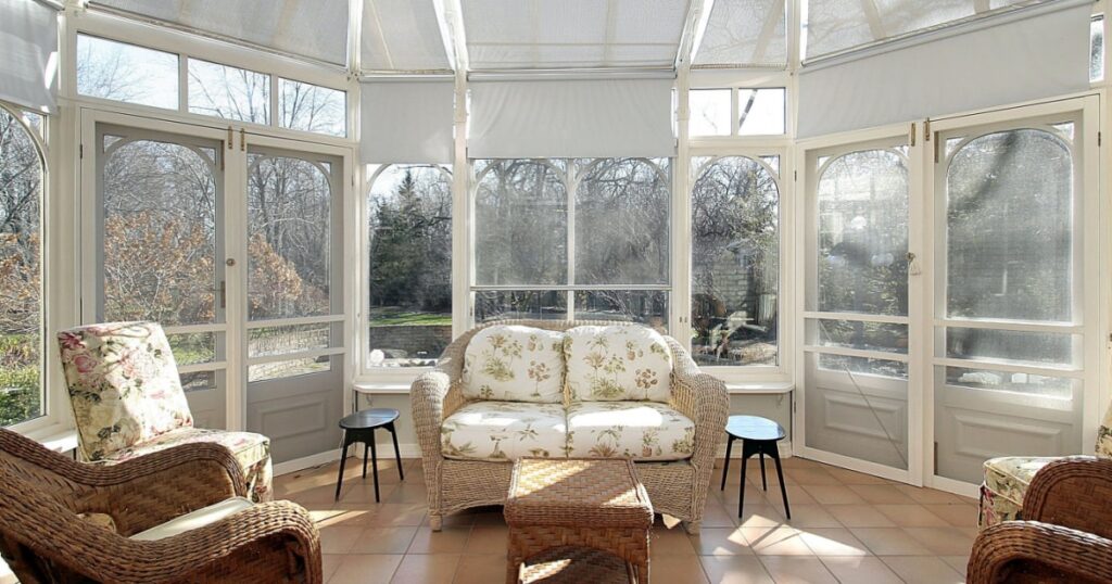 Vue intérieure d'une grande véranda victorienne blanche aménagée en salon avec des fauteuils et un canapé en rotin et tissu fleuri. 