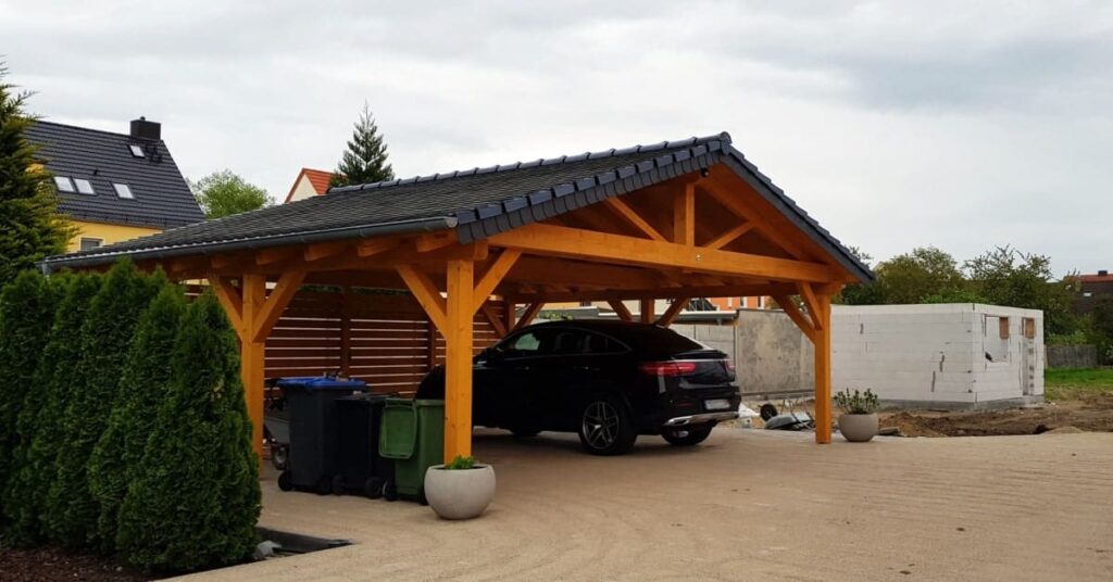 Carport en bois avec une toiture en double pente recouverte de tuiles ardoises et qui abrite une voiture. 