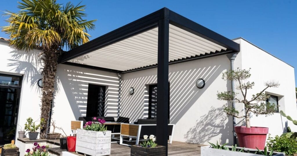 Une pergola noire en aluminium aux lames orientables adossée à une maison et abritant un salon de jardin. 