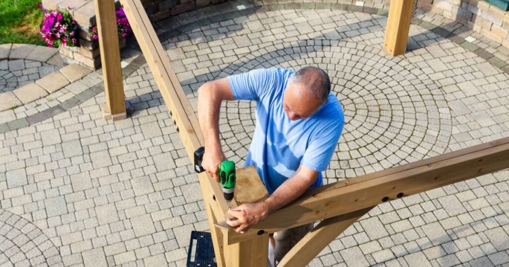 Un homme assemble sa propre pergola en bois à l'aide d'une perceuse. 