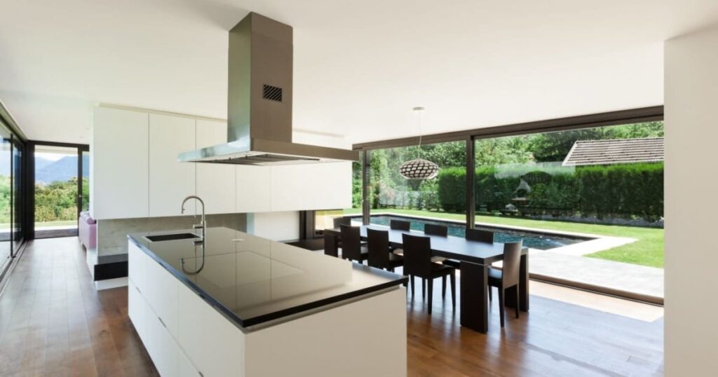 Vue intérieure d'une grande véranda avec vue sur un jardin et une piscine, aménagée en cuisine moderne avec îlot central.